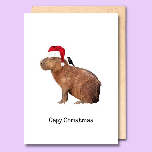 Greeting card with an image of a capybara wearing a santa hat on the front. There is a bird sitting on its back. The text says “Capy Christmas.”