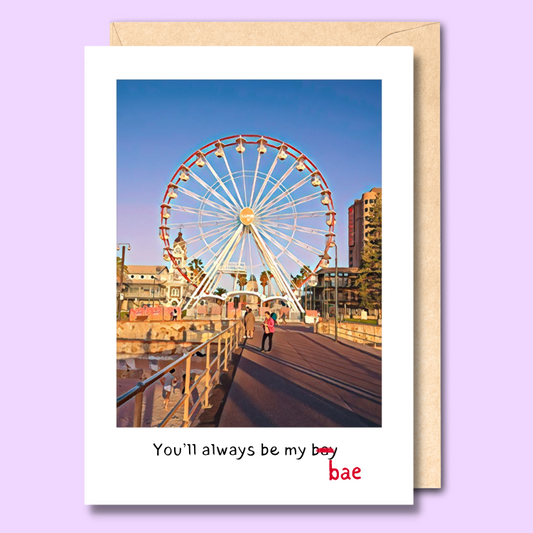 Greeting card with a photo of Glenelg jetty with a ferris wheel on the front. the text below the image says "you'll always be my bay". The word bay has been crossed out and replaced with the word 'bae'.